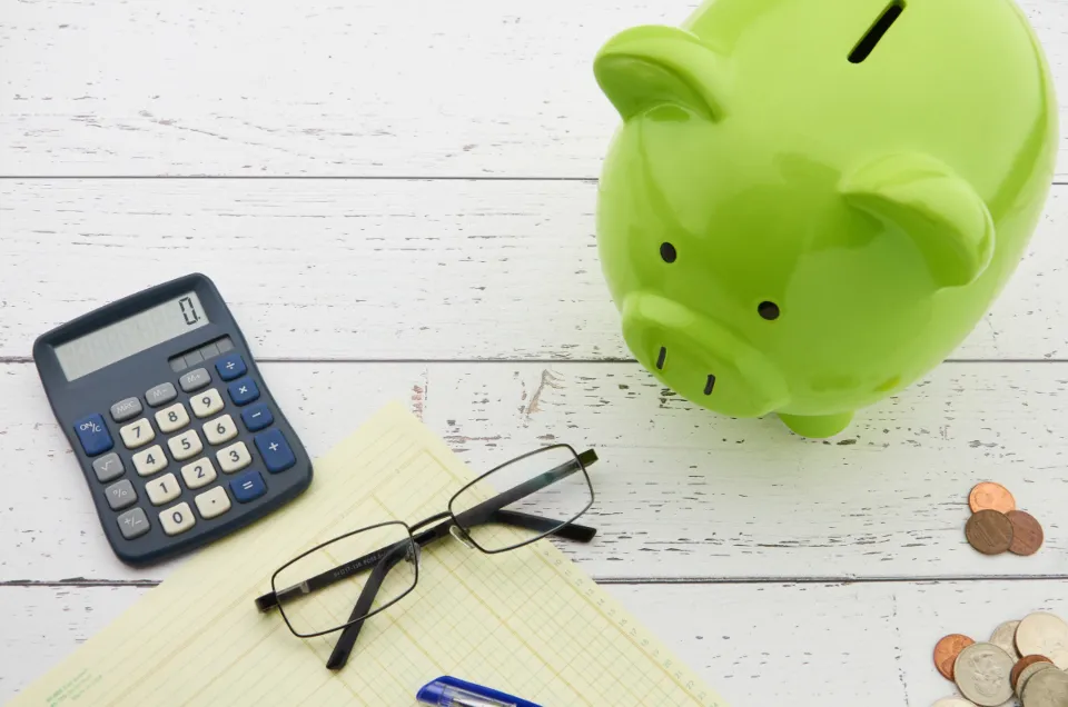 Green piggy bank next to a calculator, glasses and coins